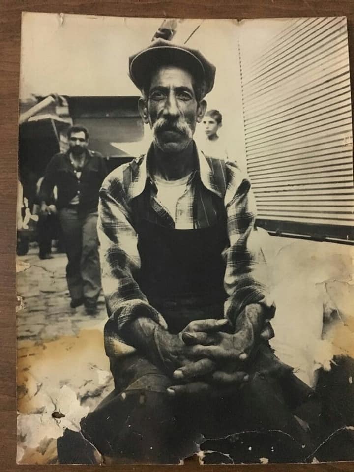 Bir yakın zaman hikayesi… Kapı Avdo. Kapı Avdo, her sabah erkenden bir başına kaldığı evden çıkar; köhne, eski dükkanının neredeyse çürümüş tahta kepeneklerini açar, çocukları gibi sevdiği kedilerini dışarı çıkararak güne başlardı. Yavru kediler de Kapı Avdo’nun gelişine sevinir, hep bir ağızdan miyavlayarak ayaklarına sokulur, kucağına atlamak isterlerdi. 60 yaşlarında olmasına rağmen hiç evlenmeyen Kapı Avdo, evden getirmiş olduğu peyniri, fırından aldığı sıcak ekmeği yemlik olarak kullandığı eskimiş aleminyum tabağa müthiş bir yavaşlıkla doğrayarak, üzerine de biraz süt dökerek, kedilerin karınlarını doyurur, kendisine demli bir çay söyleyerek,ekmek peynirle azıcık kahvaltı yaptıktan sonra da dükkanında ki işlere koyulurdu. Yemeniciydi Kapı Avdo. Dar ve karanlık , toprak damlı dükkanında gece gündüz yemeni yapar, şipariş üzerine yaptığı kösele yemenilerden, deri ayakabılardan elde ettiği gelirle yaşamaya çalışırdı.Her ayakkabıyı tek tek eliyle kalıplara alır,diker ve giyime hazır hale getirirdi. Kuri Aşiretinden olan ve Viranşehir’den geldiği söylenilen Kapı Avdo, uzun ve ince boylu, kırlaşmış pala bıyıkları, çökmüş yüzüne rağmen müthiş bir karizması vardı Siverek’te. Uzun boylu olduğu için de Kapı Abdo denilirdi ama asıl adı Abdo’yê Nazo’ydu.Nenesinin adıyla anılmasına rağmen, heybetli cüssesi nedediyle Kapı Avdo olarak bilinirdi.Az konuşan, kimilerince esrarkeş olarak da tanınan bu yaşlı amca, kelli felli birisi olmamasına rağmen Siverek’te en çok tanınan insanlar arasındaydı. Kimdi, hangi aşirettendi bilen pek yoktu ya da kimse bu yönüyle tanımazdı onu. O, kendi başınaydı. Ne kardeşleri, ne aşireti, ne de ailesi sayesinde tanınıyordu. Siverek’in Şeytan Küçesinde, Hec Abzer Hanı girişinde köhne bir dükkanda, deri kösele kundura dikerek yaşamını sürdürür, onlarca kediye sahiplik eder, onlara bakardı. Hem de evlatları gibi. Yaz –kış omuzlarından indirmediği ama hiçbir zaman da kolunu geçirmediği gabardin ceketi hep omzundaydı. Yılların solmuşluğunu taşısa da o ceketini değiştirmezdi. Suriye’den hediye gelen ceketin altına yine gabardin kahverengi şalvarı giyer, kendi elleriyle yaptığı kösele kunduraların topuklarını kırarak ve ellerini arkasında bağlayarak her sabah Kale Boğazı’ndan ağır ağır yürüyerek ve sıklıkla derin derin öksürerek dükkanına gelirdi. Dükkanına geldiğinde ilk iş kedilerini doyurur, yıllardır boyanın ve yapıştırıcının etkisiyle tahtalaşan önlüğünü giyer, elindeki “ drevti” denilen deliciyi bir deriye, bir köseleye batırarak kundura yapmaya başlardı. Kösele yumuşasın diye birkaç gün suda bekletir, delicisini sürekli sivrilterek işinin çabuk olmasını sağlardı. Kuzu derisi kullandığı ayakkabılar özellikle orta yaş insanlarda oldukça rağbet görür, herkes Kapı Avdo’nun ustalığına vurgu yapardı. Ağaları, beyleri , zorba insanları sevmezdi Kapı Avdo… Arkadaşlarının çoğu yoksul ve meteliksiz insanlardı. Delileri çevresinde toplar, ekmeğini ve sigarasını onlarla paylaşırdı. Gelen misafirlerine çay ikram eder, köhne de olsa dükkanının bir kenarında oturturdu. En çokta “ talebe” dostları vardı. Okuyanları sever, sayardı. Sabahın köründe başladığı çalışmasını yazın sıcak bastırana kadar sürdürür, sonra ara verir, önlüğünü çıkartır, dükkanını öylece bırakıp, ceketini omuzlarına alarak Siverek’in dar sokaklarına dalardı. Kapı Avdo istisnasız her gün bunu yapar ama nereye, niçin gittiğini bilen olmazdı. Soran olsa da cevap vermez,sigarasından derin derin duman çekerek, yoluna devam ederdi. Dükkanı ise öylece açık kalır, her şey yerli yerinde dururdu. Kapı Avdo yıllarını böyle geçirdi. Parmaklarındaki dırevti yaraları nasırlaştı, nasırlar kemikleşti yıllar içerisinde. Ekmeğini kedilerle paylaştı. Devrimcilerin dünyalarına yakın durdu, dükkanında bildirilerini sakladı,Yılmaz Güney’in posterlerini toprak sıvalı dükkanının en aydınlık yerine astı… Onun heybetli boyu ve karizmatik kırlaşmış bıyıkları Şeyh Bedrettin’i andırdığı için o yıllarda Siverek ve çevresinde belgesel çekmeye gelen Ünlü Fotoğraf Sanatçısı Tuğrul Çakar’ın dikkatini çekmiş, dükkanının önünde ki tezgah başında ki hali fotoğraflanarak ölümsüzleştirilmişti. Siverek’in en kadim ayakkabı ustası Kapı Avdo’nun bu olağanüstü siyah beyaz fotoğrafı aynı zamanda bir kartpostala dönüştü. Nazım Hikmet’in Şeyh Bedrettin adlı şiirinin dizeleri Kapı Avdo’nun heybetiyle birleşince, ortaya sahici bir Şeyh Bedrettin sülieti çıktı. Kartpostal kısa sürede en popüler kartların arasına girmeyi başardı, elden ele dolaştı. Ama kimse fotoğraftaki kişinin Kapı Avdo olduğunu bilemedi. O da kartpostallardaki fotoğrafına aldırış bile etmeyerek yaşamına devam etti.Yıllarca sol düşünceyi taşıyanlar bu kartpostalı cezaevlerine, sevdiklerine, dünyanın diğer uçuna gönderdi. Şeyh Bedrettin yıllar sonra Kapı Avdo’nun köhne dükkanında yeniden milyonlara ulaştı. O yıllarda (1978-79-80) Siverek ateş çemberinden geçti, gençler dar ağacına, işkence tezgahalarına alındı. Her şey hızlıca darmadağın olurken, Kapı Avdo yaşam biçimini hiç değiştirmedi. Az konuşan ama konuştuğunda da çok şey söyleyen birisi olarak yalnızlığı sürdürdü. Çok misafiri olsa da, o hep bir başına yaşadı. Kapı Avdo’nun bir gün dükkanı açılmadı. Hayatında ilk kez dükkanını açmıyordu. Siverek’in dışına bile çıktığı duyulmamış, görülmemişti.Bir yere gitse, anahtarı birilerine verir, dükkanını açtırırdı. Ama bu kez dükkan açılmamıştı. İçerde mahsur kalan kediler o gün akşama kadar miyavladılar, kepenekleri tırmaladılar. Komşuları kötüye yormak istemediler Kapı Avdo’nun gelemeyişini. Ertesi gün kara haber ulaştı Şeytan Küçesine. Kapı Avdo tek başına kaldığı toprak damlı evde, Ahmed Arif’in şiirlerinde yazdığı gibi bir kuşluk vakti ölmüştü, habersiz ve kimsesiz. Sıvası dökülmüş duvarlar, birkaç parça kap kacak ve Suriye malı ceketi odanın orta yerindeydi. Anlaşılan kendisini zor atmıştı yer döşeğine. Ölüm onu bir başına yakalamıştı. Zulalarında birkaç adres ve çok az bir para bulundu. Kurumuş bir ekmek, yarım bırakılmış bir sarılı kalın sigara ve Halep malı bir tütün tabağı Kapı Avdo’nun özel eşyaları olarak toplandı… Yalnızdı, yaşlı ve kimsesizdi Kapı Avdo. Akrabaları, kardeşleri vardı ama o bir başına yaşamayı tercih etmişti. Öldüğünü duyan deliler, kimsesizler o gün ağladılar durmadan. Kedileri miyavlayıp durdu kepeneklerin arkasından. Anlamışlardı sanki kötü bir şey olduğunu. Ertesi gün komşular kepenkleri açar açmaz kedilerin her biri bir tarafa kaçıştı. Kapı Avdo’yu aradılar insan kalabalıkları arasında. Ama Kapı Avdo yoktu, geriye dönülmeyecek bir yolculuktaydı. Sokaklarda aç aç dolaşan kediler birkaç gün sonra Kapı Avdo’nun semtini terk ettiler ve kayboldular ortalıktan. Kapı Avdo bildiği gibi yaşayan, yoksul ve bir o kadar da mağrur birisi olarak yaşamını noktaladı.Kedilerle, delilerle, devrimcilerle paylaştığı sofrası ve nasırlı elleri onu ölümsüzleştirdi Şeytan Küçe’sinde. Aradan 40 yıl geçti. Kapı Avdo’nun karanlıklar içerisinde, bütün heybeti ile oturan resmini görünce içimden bir şeyler koptu sanki. O yıllara, çocukluğumun geçtiği sokaklara geri döndüm… Hayatın siyah beyaz olduğu yıllara döndüm. Kaç insan hatırlanır ki onca yıl sonra? … Merhum Tuğrul Çakar’ın objektifinden günümüze ulaşan siyah beyaz fotoğrafı Kapı Avdo’nun hikayesinin bir özetti gibi. Yorgun gözler, kırlaşmış bıyıklar ve nasır tutmuş eller… Kapı Avdo yüreğinde kaç sevda eritti, kaç vurgun yedi bilinmez. Bilinen o ki her gün yeniden acıtarak, kanatarak yüreği ateşlerde kavruldu, hançerlendi, yaşamın girdabında çırpındı ve ansızın sessizce terk ê diyar etti..